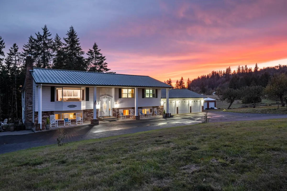 The image showcases the front facade of a spacious single-family home at sunset, with a sweeping driveway leading up to the inviting porch. The sky is painted with warm hues, casting a tranquil glow over the peaceful countryside setting. The home exudes warmth and welcome, with plenty of outdoor space ideal for gatherings and relaxation.
