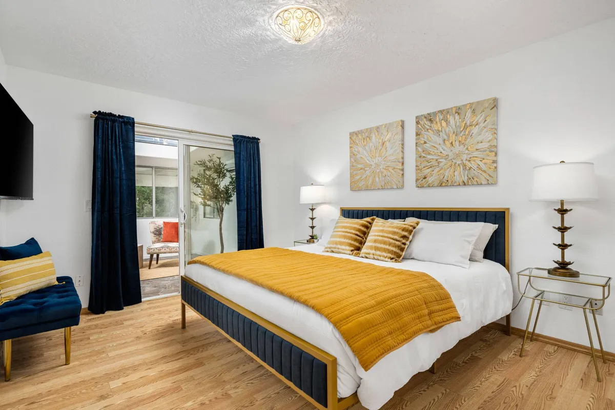 A sunlit bedroom with a large window and inviting bed, draped in a yellow blanket, with decorative wall art and a nightstand. The open window hints at natural light and the serene, peaceful setting offered by the property.
