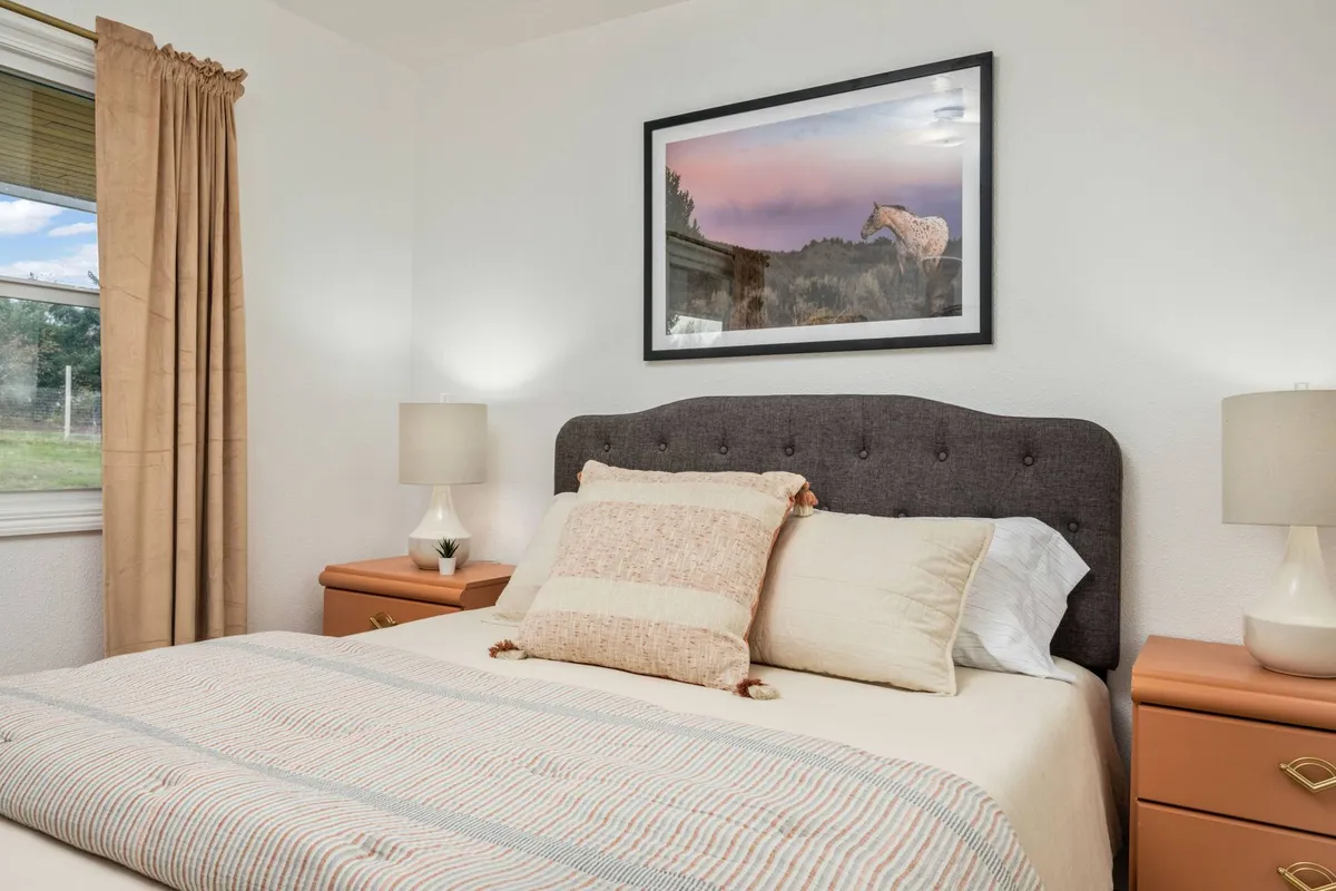 This image features a serene bedroom with a tufted headboard bed, soft bedding, framed horse artwork, and bedside lamps. The natural light filtering into the room creates a peaceful, inviting ambiance, perfect for relaxation and restful sleep.