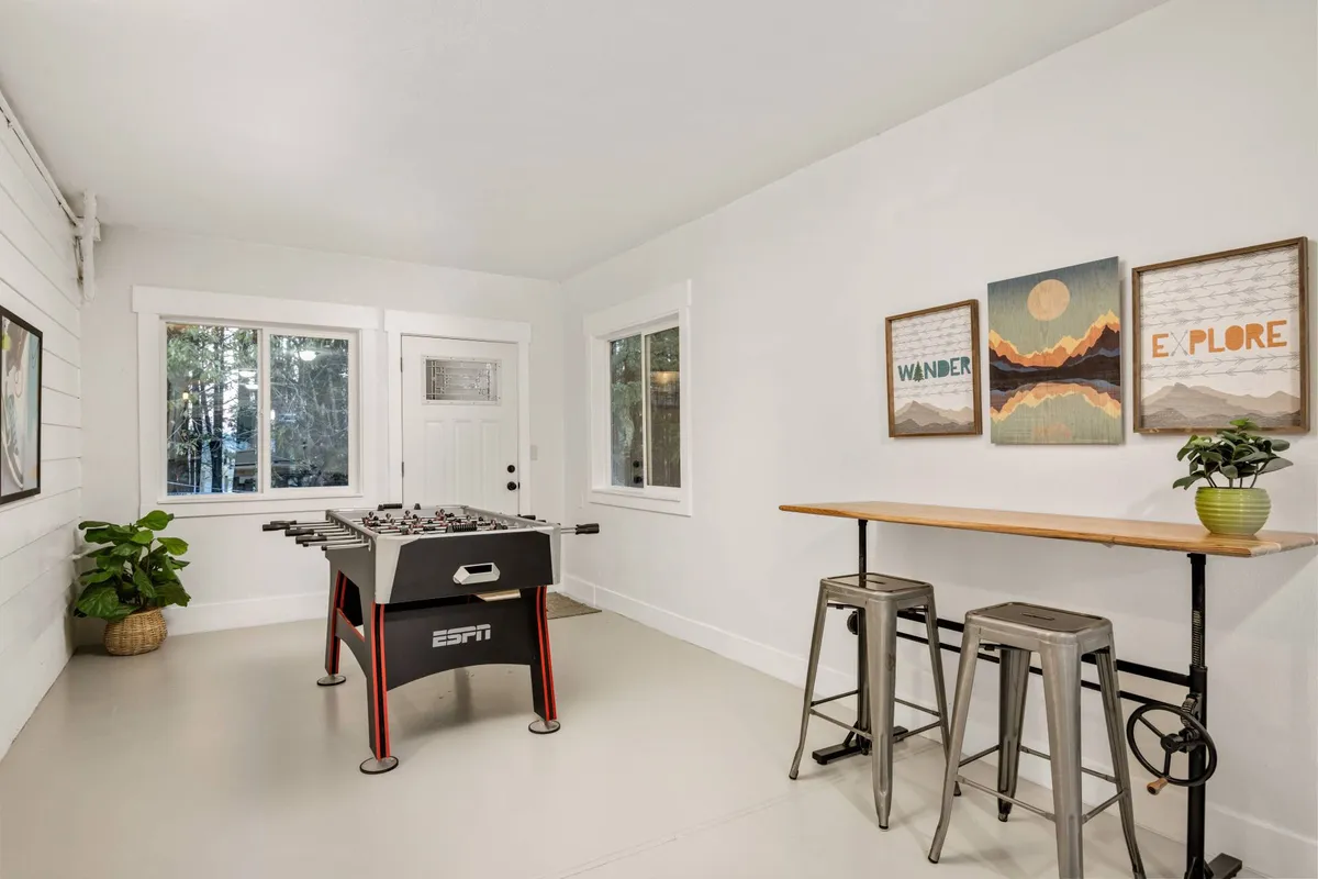 A casual game area featuring a foosball table, a bar table with stools, and motivational wall art. Large windows let in abundant natural light, and the space feels open and playful, inviting competitions and camaraderie.