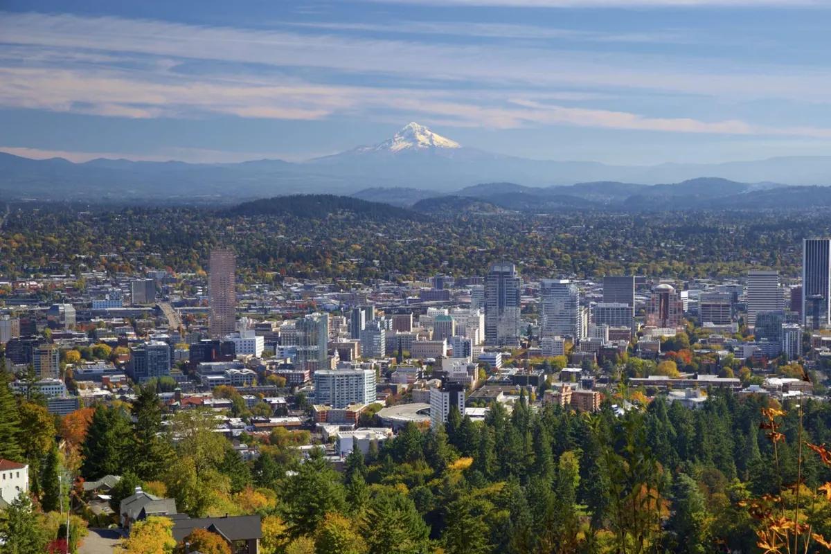 A panoramic view of the city skyline with Mount Hood rising in the distance, framed by lush greenery and vibrant fall foliage. The blend of urban and natural elements showcases both proximity to city amenities and the scenic beauty of the region.