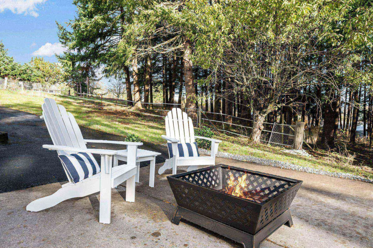 Two white Adirondack chairs face a black fire pit with a fire burning, surrounded by lush green grass and mature trees in the background. The setup invites relaxation and conversation amid nature.