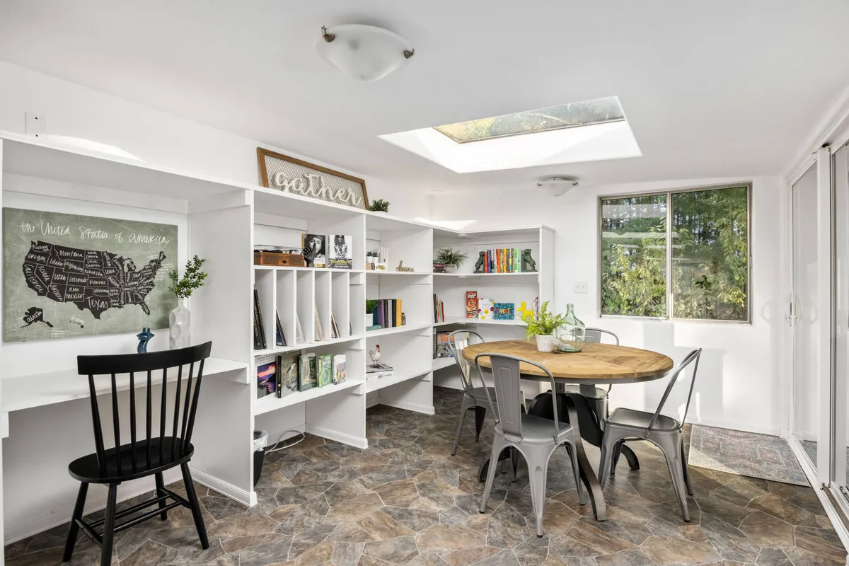 A communal dining area with a round wooden table, black and white map artwork on the wall, and open bookshelves. The space is arranged for family meals, reading, and lively game nights, fostering a sense of connection and shared experience.