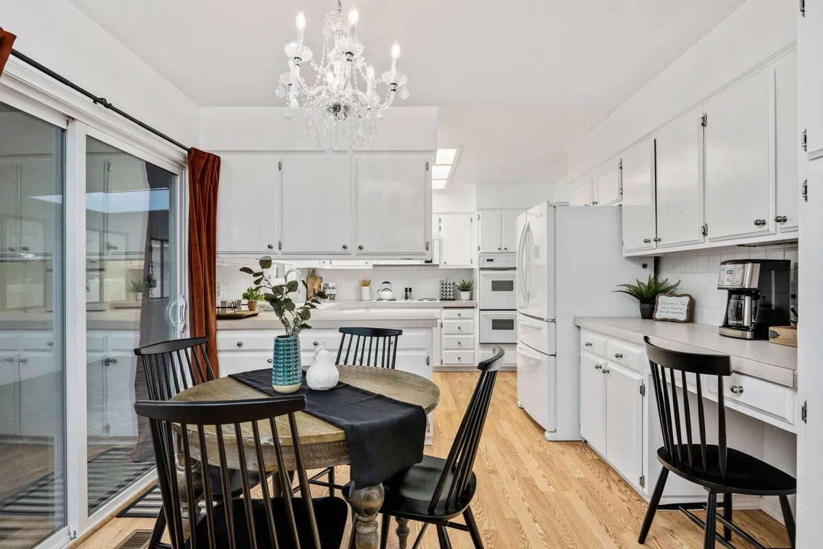 A bright open-plan space with a chandelier above a generous dining table, visible kitchen appliances, and a sliding glass door that brings in natural light. The setting is ideal for family meals, celebrations, and casual gatherings.