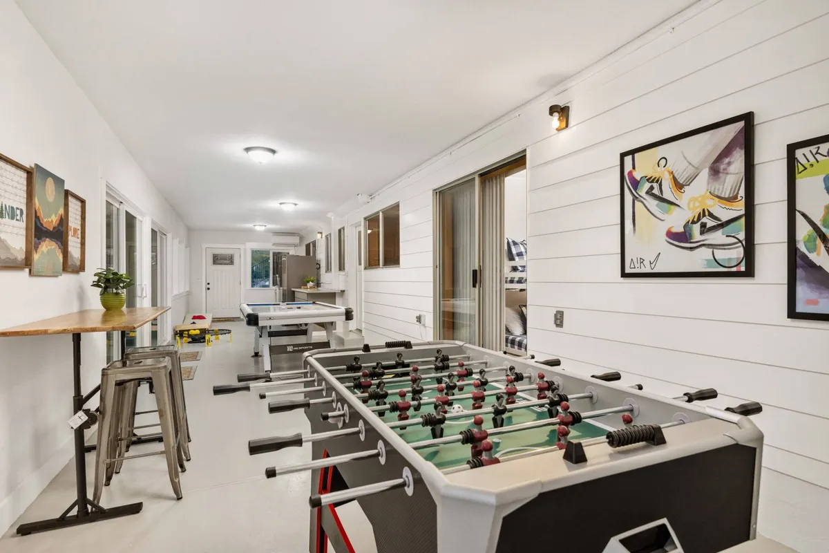 A bright and lively game room featuring a foosball table and a ping pong table, with bar-height stools and wall art adding to the energetic atmosphere. Natural light streams in through large windows, making the space inviting for socializing and friendly competition.