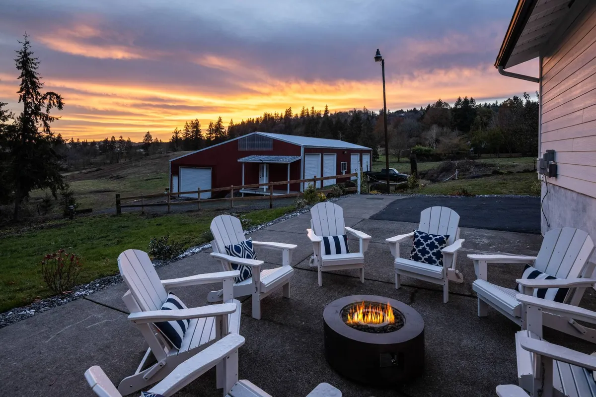 This image captures a patio scene with Adirondack chairs encircling a glowing fire pit, set against a backdrop of a rustic barn and open sunset-lit sky. The surrounding landscape hints at rolling lawns and farm fields.