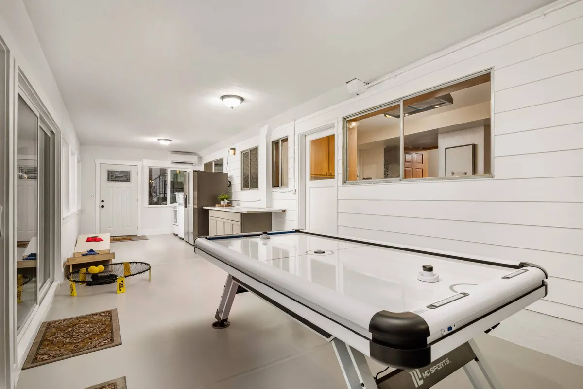 A lower-level kitchenette featuring a modern kitchenette area, air hockey table, large windows for natural light, and an entrance door—conveying a relaxed, playful secondary gathering space ideal for group living.