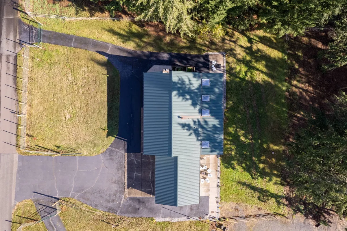 A scene showing the house with a green metal roof, surrounded by abundant greenery and a paved driveway. The image evokes a sense of tranquility, privacy, and accessibility, with natural beauty and ample parking visible.